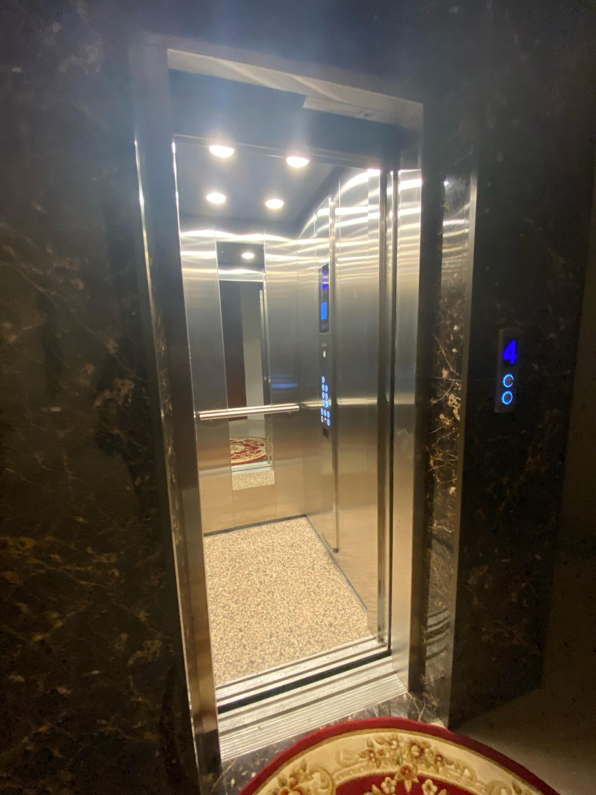 L’intérieur d’un ascenseur, avec un sol en marbre, un écran numérique et une zone recouverte de moquette.