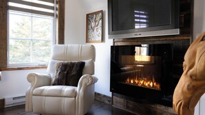 A living room with a fireplace, a white leather armchair, a television, and a wooden head sculpture. - La Ruelle Hébergement (Saint-Siméon-de-Bonaventure)