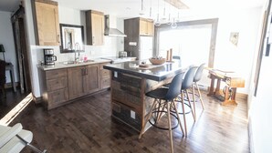 A modern kitchen with wooden cabinets, a central island with bar stools, and a large window providing natural light. - La Ruelle Hébergement (Saint-Siméon-de-Bonaventure)