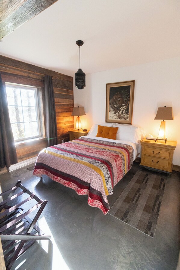 A bedroom with a large bed, two bedside tables with lamps, a wooden headboard, a window with curtains, and a painting on the wall. - La Ruelle Hébergement (Saint-Siméon-de-Bonaventure)
