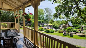 A covered outdoor area with a wooden deck, a table with chairs, and hanging flower baskets. - La Ruelle Hébergement (Saint-Siméon-de-Bonaventure)