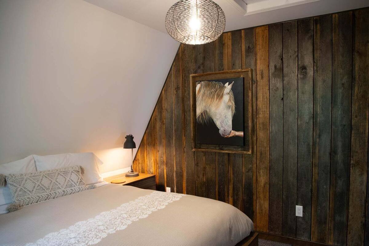 A bedroom with a wooden wall, a bed with white bedding, a lamp, and a framed picture of a horse.