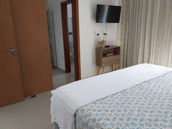 A bedroom with a bed, a TV mounted on the wall, a wooden nightstand, and a door leading to another room.
