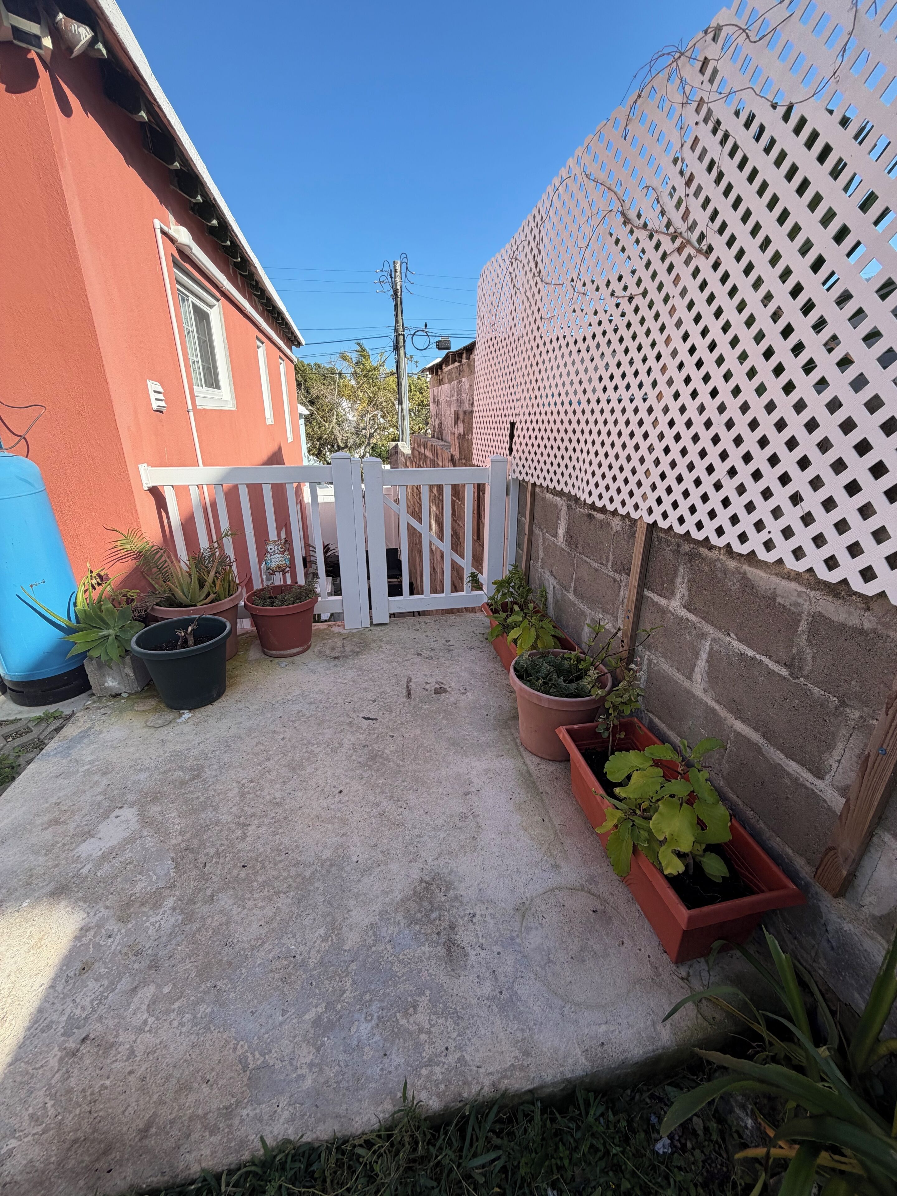 Un patio de concreto con plantas en macetas, una cerca blanca de piquetes y un edificio rojo.