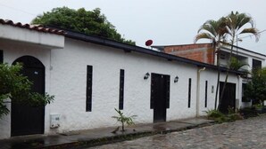 A white building with black doors and windows, a stone-paved street, and a palm tree. - Cabaña el árbol - Quadruple Room (Jagua)