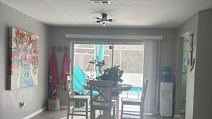 A spacious living room with a ceiling fan, a dining area with a round table and chairs, and a large window with a view of the outdoors. - Beachy themed beautiful home with private pool available May thru Nov! (Lehigh Acres)