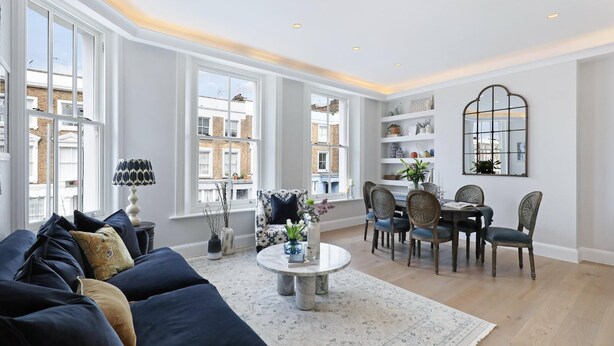Un salón amplio con un sofá grande, una mesa de comedor con sillas y una mesa auxiliar. Hay ventanas grandes que ofrecen luz natural y vistas de la ciudad.