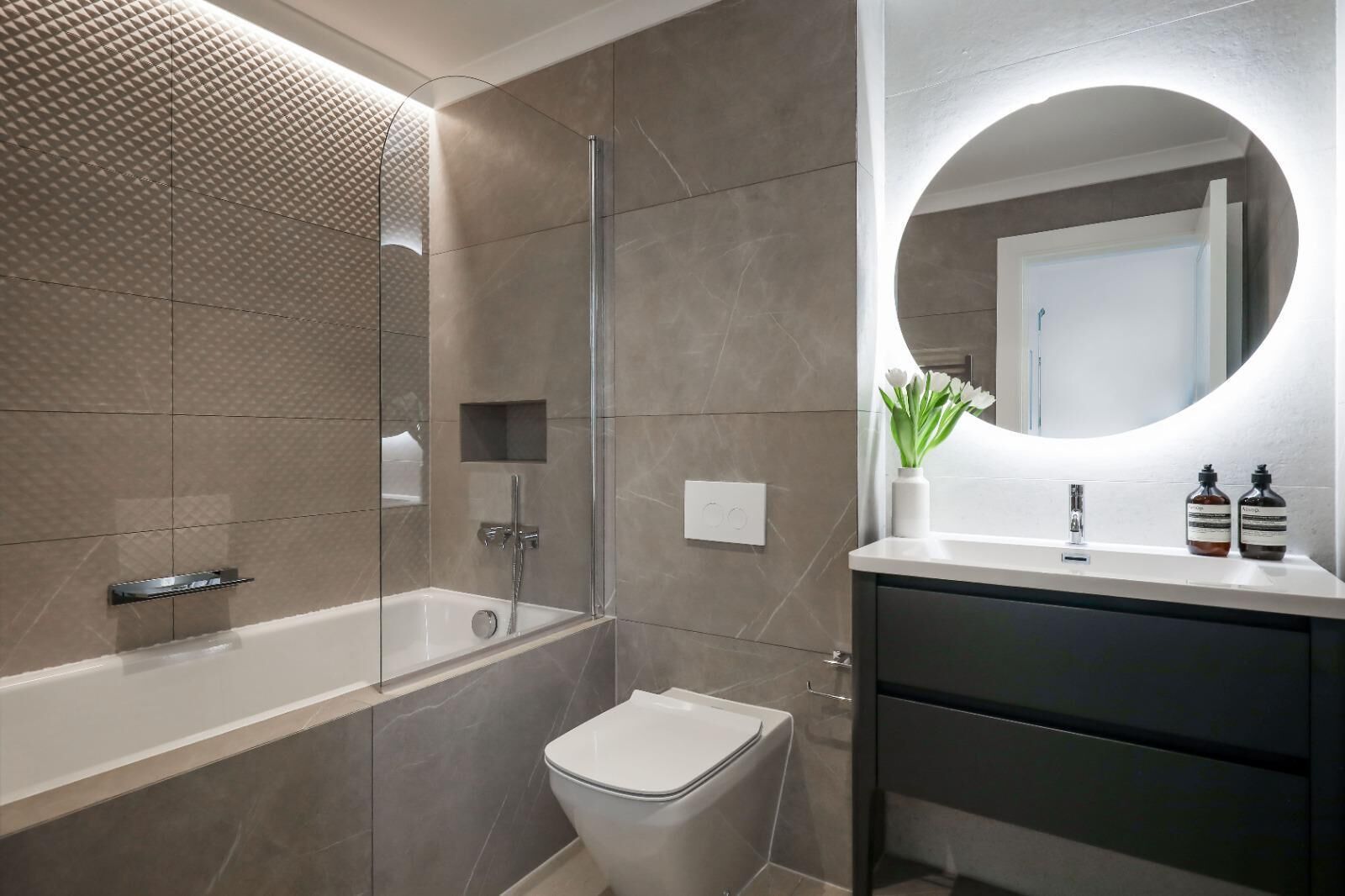 A modern bathroom with a glass-enclosed shower, a white toilet, and a vanity with a mirror and two bottles.