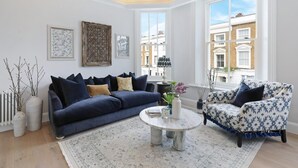 Una sala moderna con un sofá azul oscuro, un sillón estampado, una mesa redonda de centro y un ventanal con vistas a edificios.