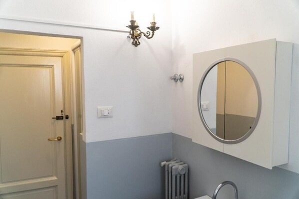 Une salle de bain avec une porte blanche, un miroir rond, un radiateur et un luminaire fixé au mur.