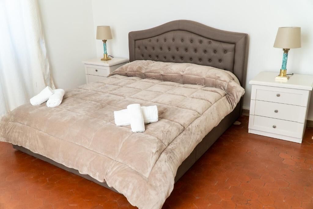A neatly made bed with a beige duvet and pillows, two bedside tables with lamps, and a tiled floor.