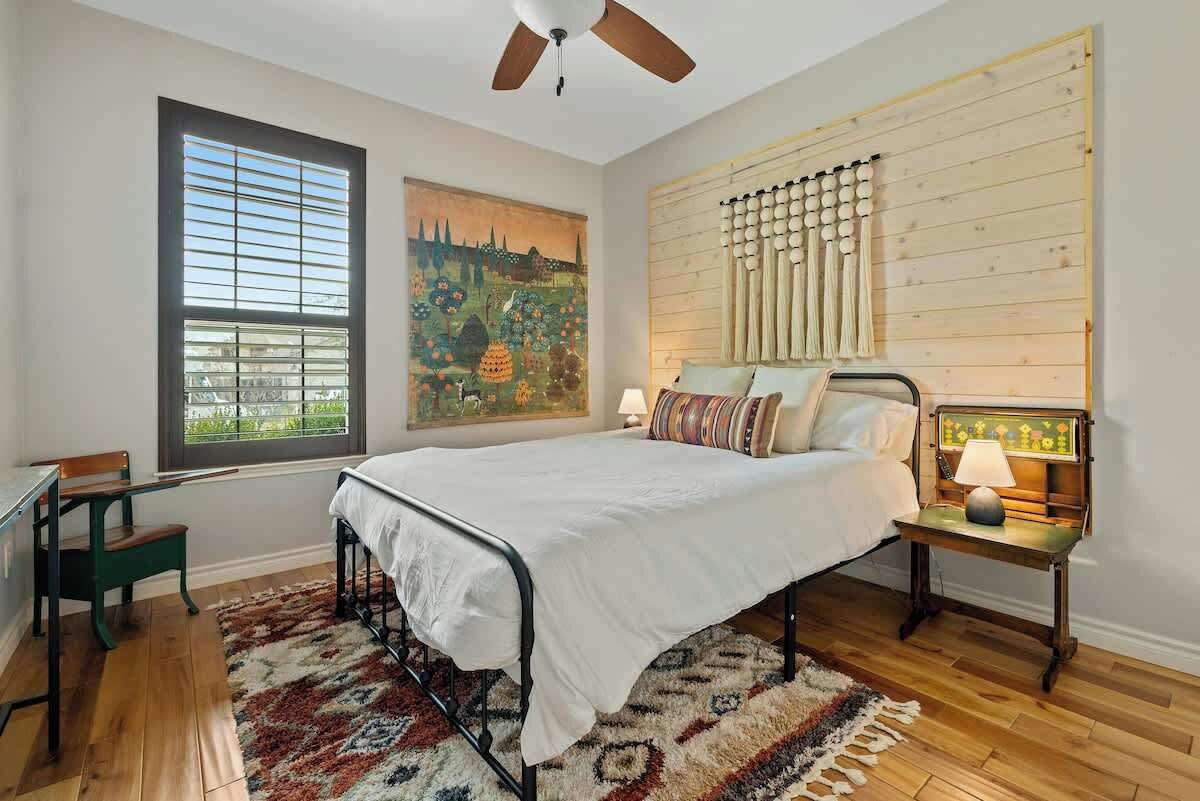 A bedroom with a bed, a desk, a chair, a window with blinds, a ceiling fan, and a wall-mounted lamp.