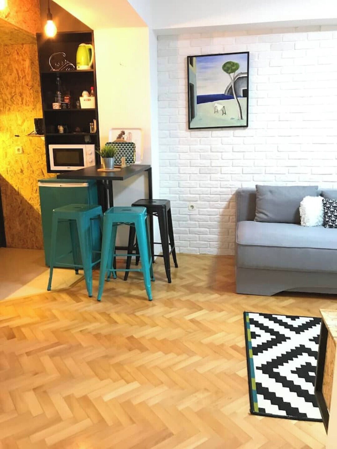 Een ruimtelijk ingerichte leefruimte met kitchenette, een grijze bank en een visgraatvloer van hout.