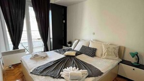 Un lit bien fait, avec une couverture et des serviettes, une lampe de chevet et une vue sur l’extérieur par une fenêtre munie de rideaux.