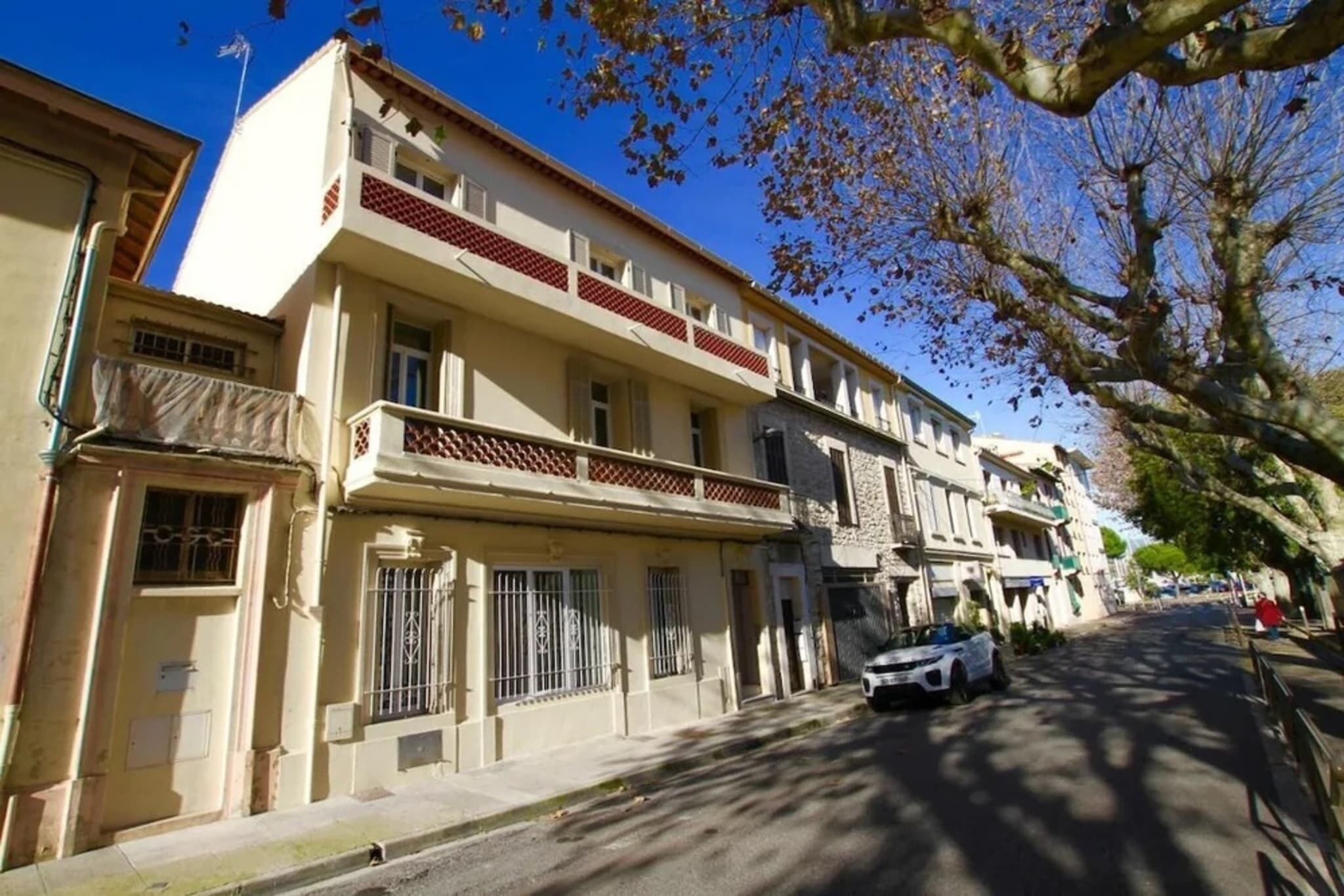 Un immeuble résidentiel à plusieurs étages doté d’un balcon, une voiture garée dans la rue et des arbres aux feuilles d’automne.