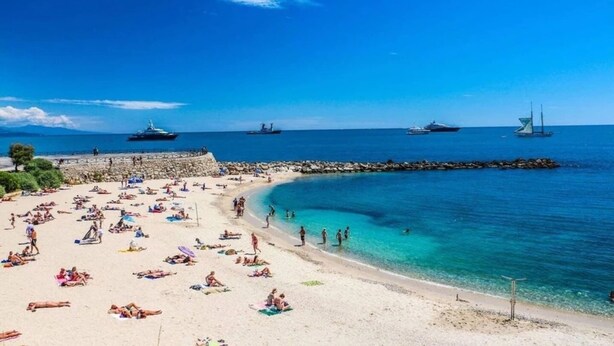 Ein Strand mit badenden Personen, einer Steinmole und mehreren Booten im Wasser.
