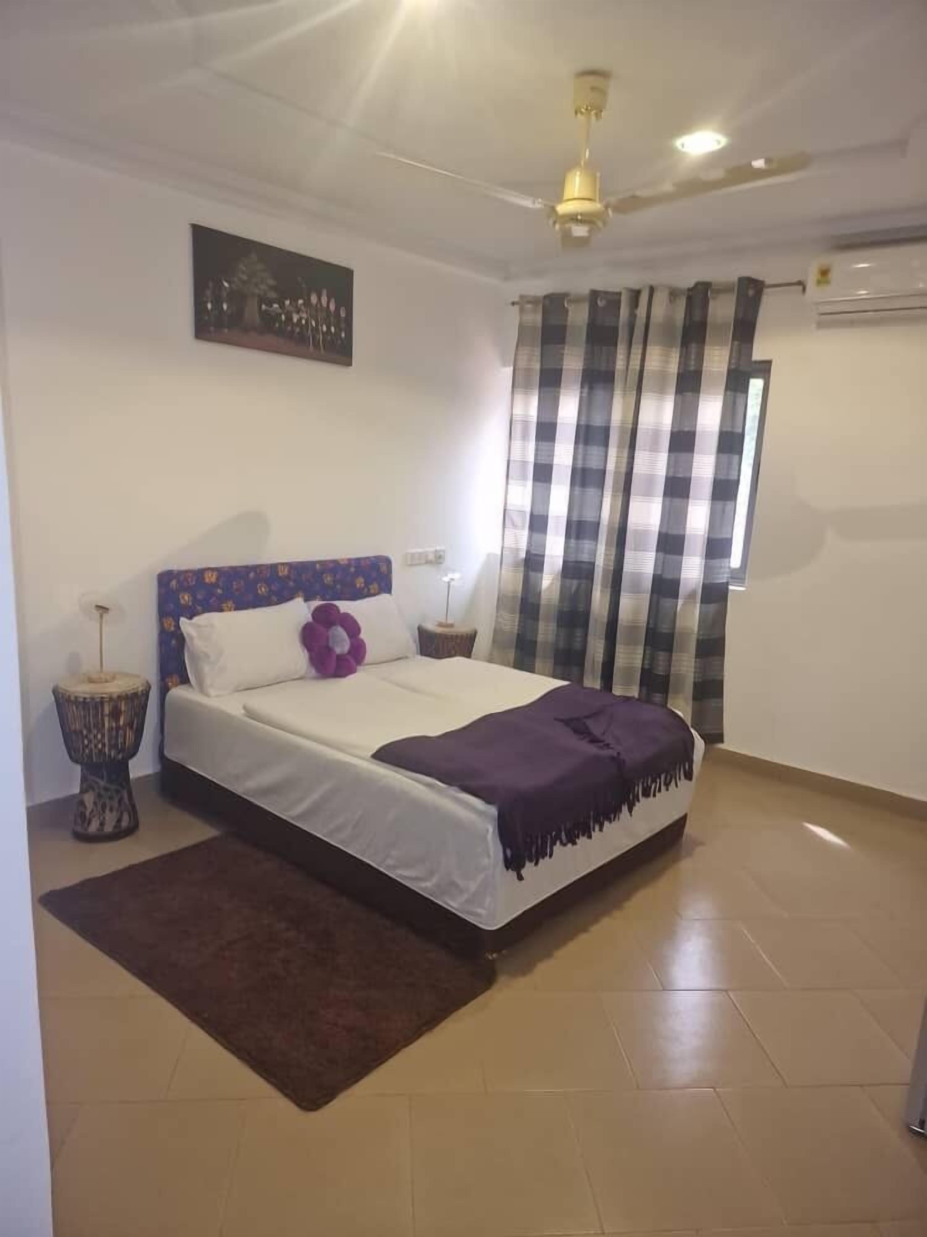 Un dormitorio con cama, ventilador de techo, ventana con cortinas, aire acondicionado de pared y un adorno en la mesita de noche.