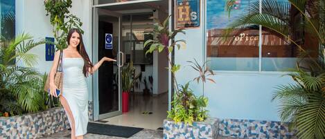 A woman in a white dress standing at the entrance of a building with a tiled floor and potted plants.