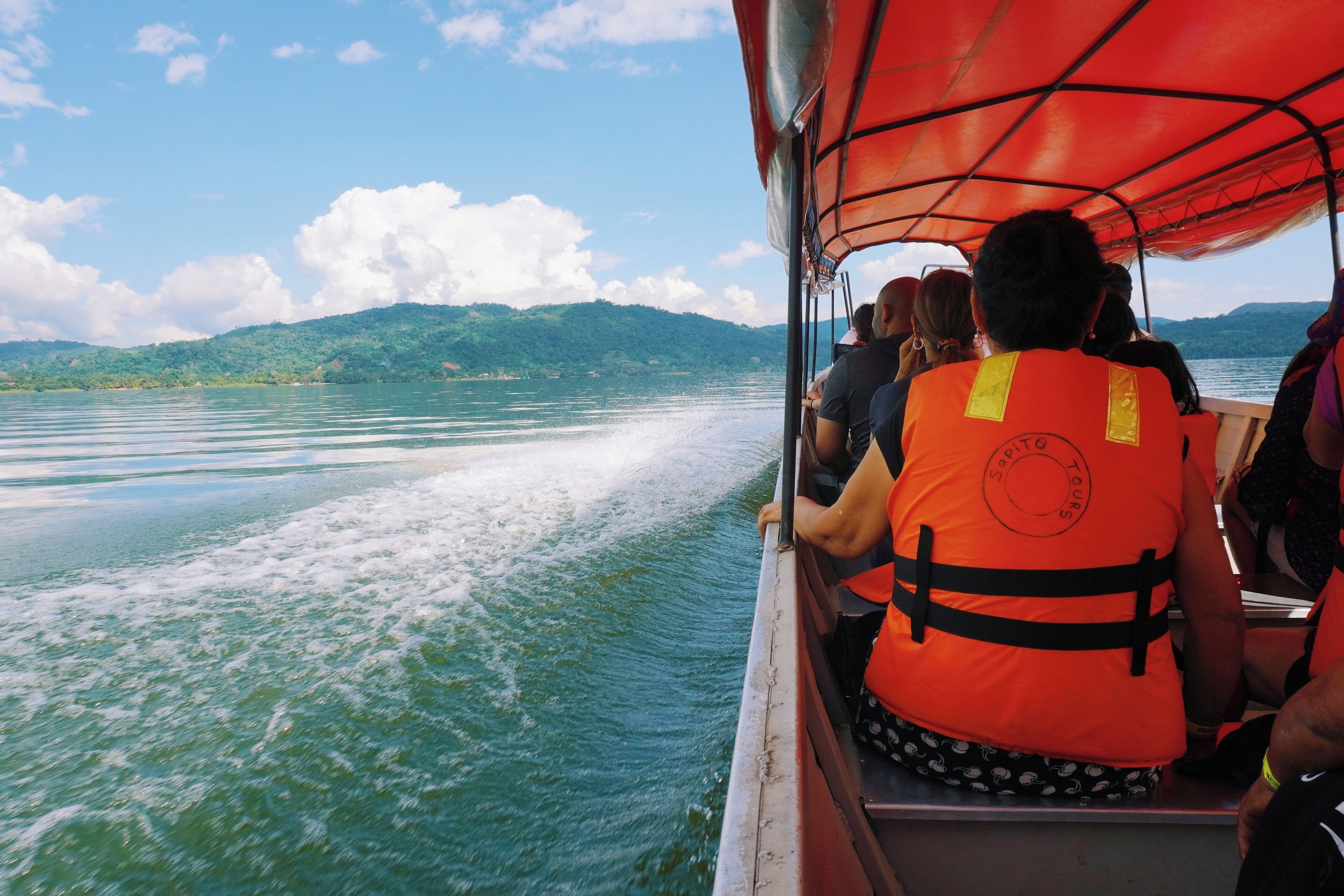 A boat with passengers wearing life jackets, moving on a calm body of water with mountains in the background.