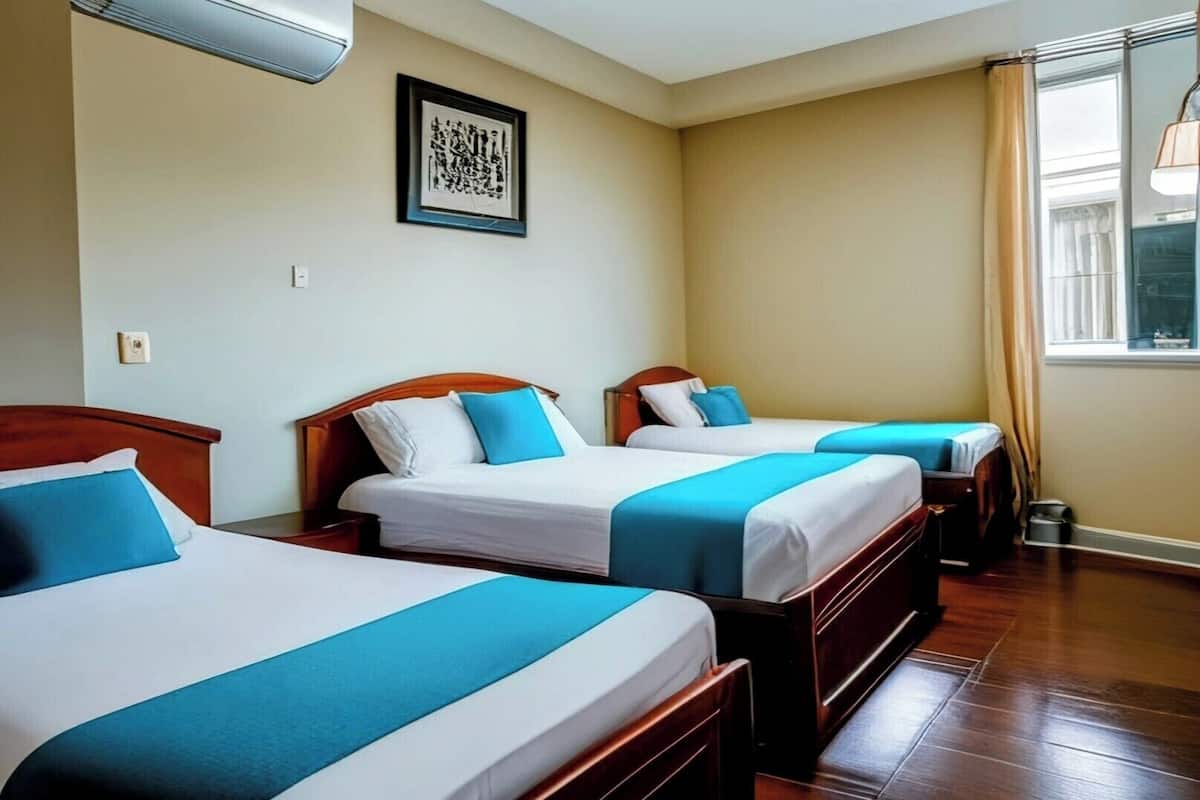 Una habitación de hotel con tres camas, cada una con cabecero de madera y ropa de cama azul. Hay una obra de arte enmarcada en la pared y una ventana con cortinas.