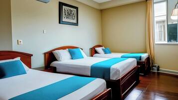 Quarto de hotel com três camas, cada uma com cabeceira de madeira e roupa de cama azul. Há uma obra de arte emoldurada na parede e uma janela com cortinas.
