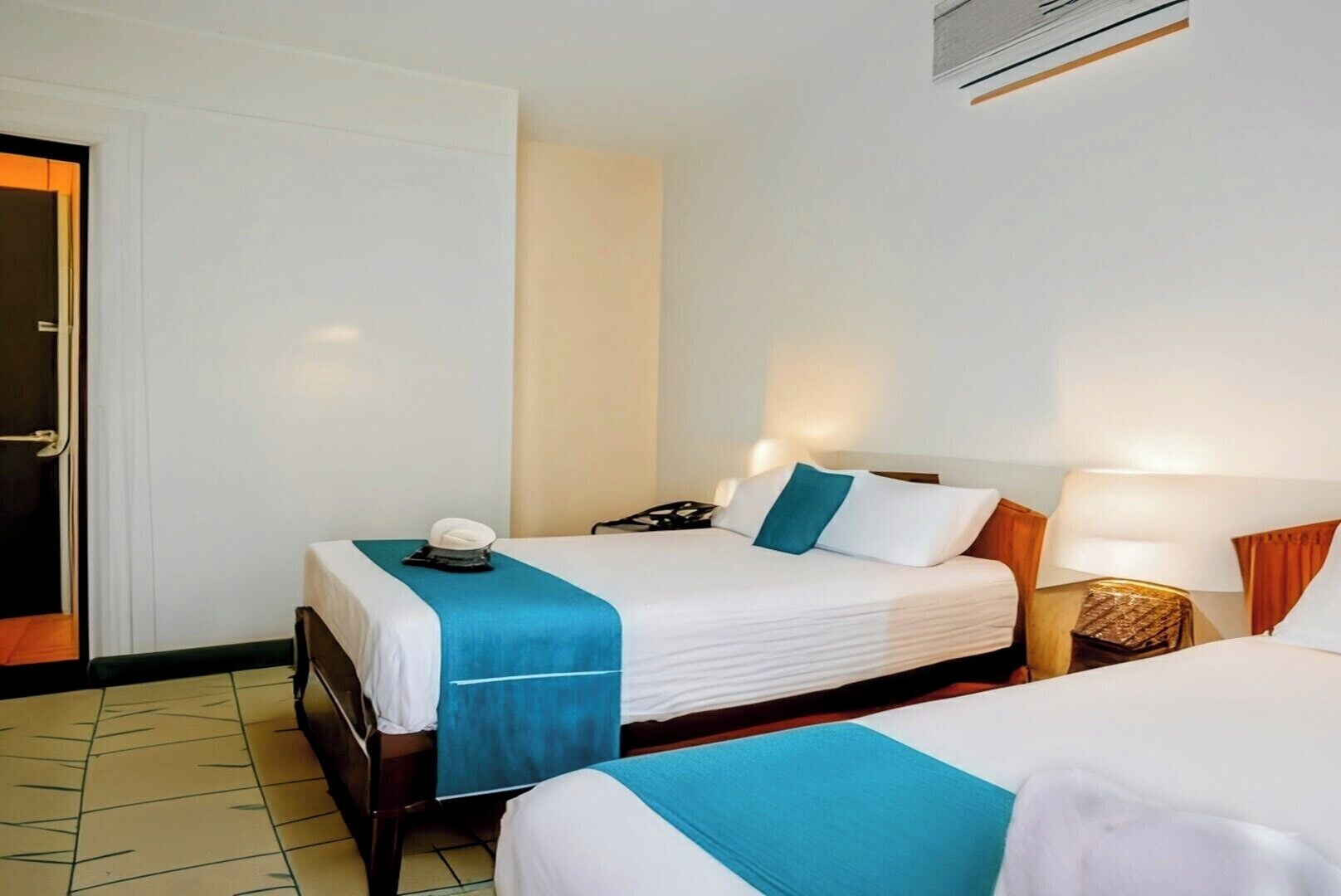A hotel room with two beds, a door, a wall-mounted air conditioner, and a bedside table with a lamp.