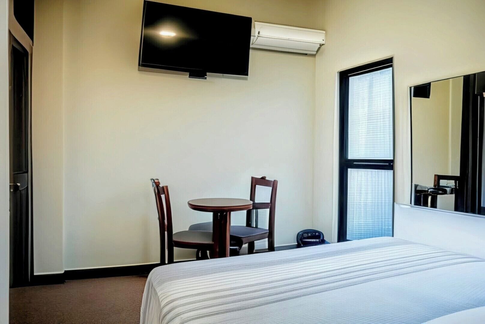 A hotel room with a bed, a small table with two chairs, a flat-screen TV mounted on the wall, and a window with blinds.
