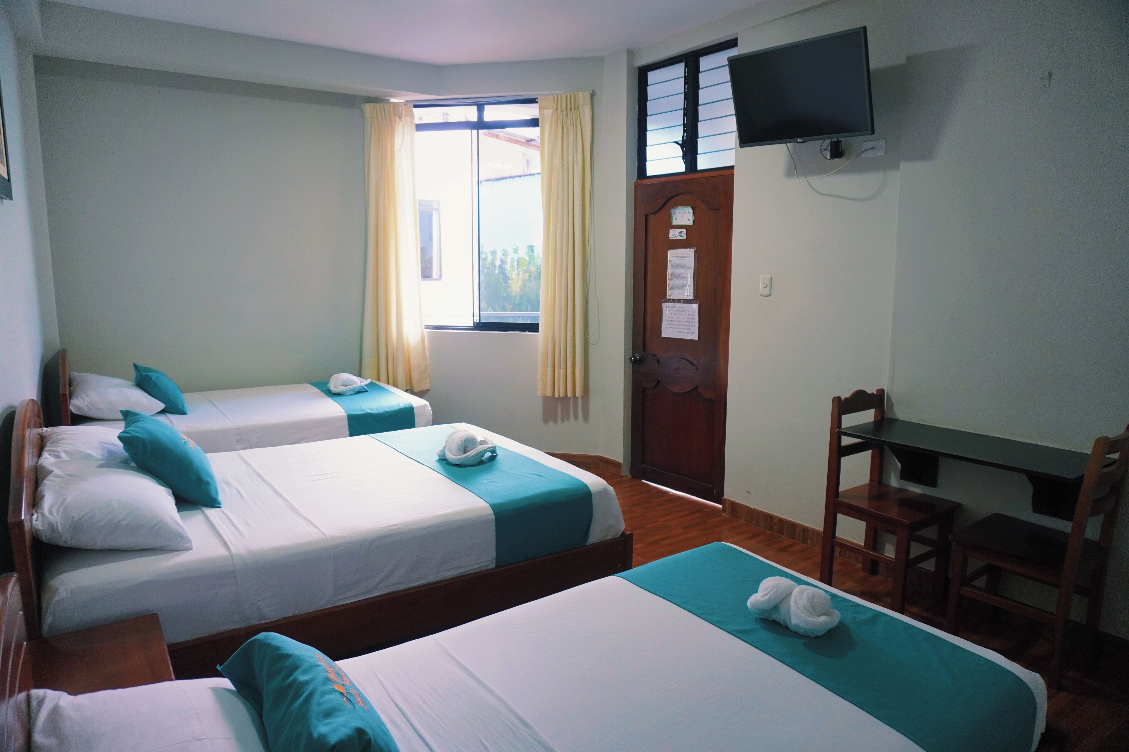 A hotel room with three single beds, a desk, a chair, a TV mounted on the wall, and a window with curtains.