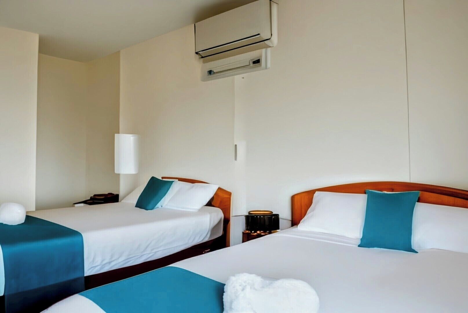 Een hotelkamer met twee bedden, elk voorzien van witte lakens en teal kussens. Er staat een nachtkastje met een lamp en een klein tafeltje met een kaars.