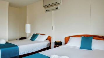 A hotel room with two beds, each with white linens and teal pillows. There is a bedside table with a lamp and a small table with a candle.