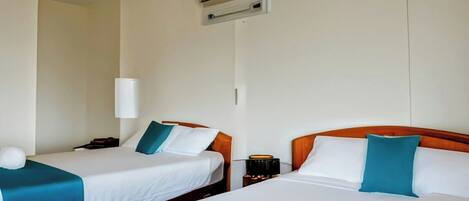 A hotel room with two beds, each with white linens and teal pillows. There is a bedside table with a lamp and a small table with a candle.