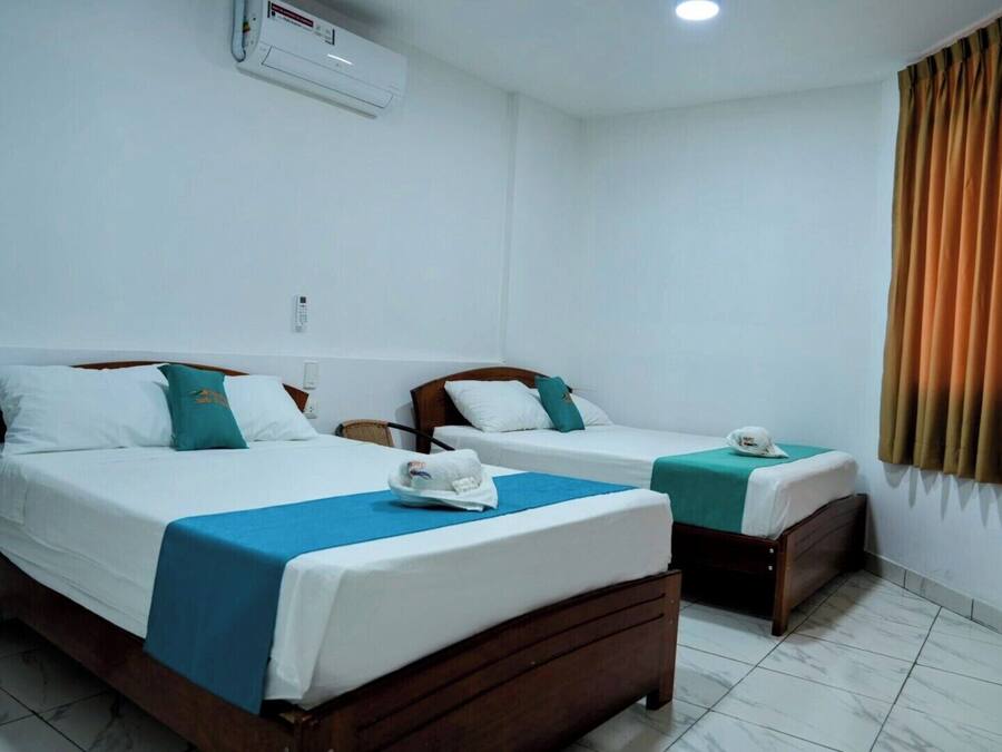 A hotel room with two beds, each with a wooden frame and white bedding. There is a ceiling air conditioner and a window with curtains.