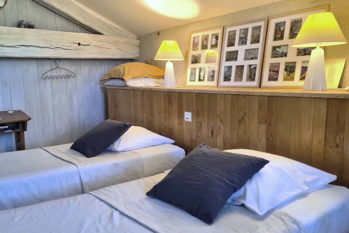 Two single beds with white linens and dark pillows, a wooden headboard, and a shelf with framed pictures and two lamps.
