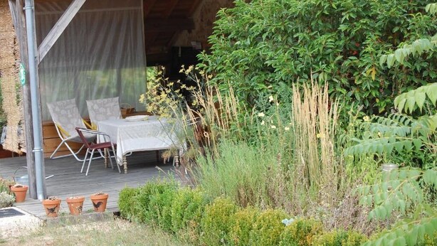 An outdoor seating area with a table and chairs, surrounded by lush greenery and a wooden deck. - Clos de l'ARTHONNET (Flavignac)