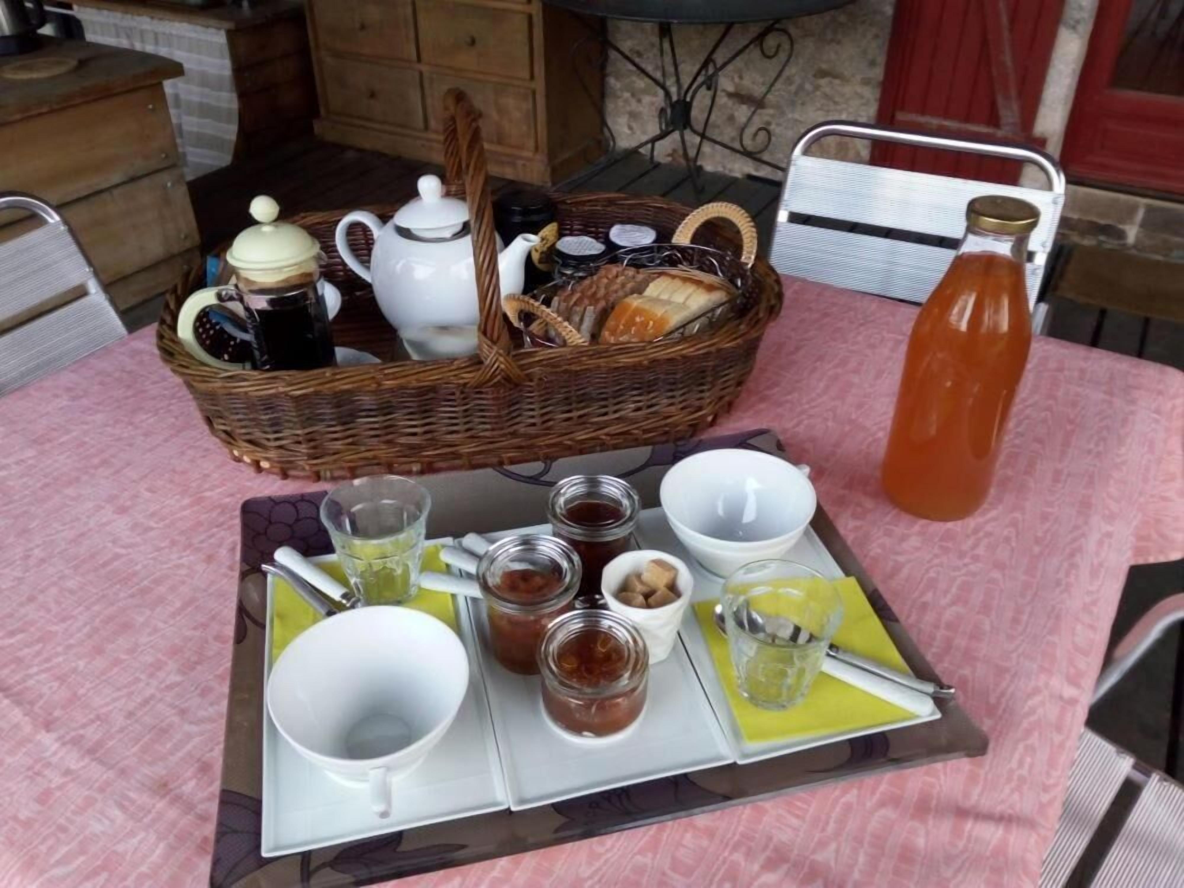 A breakfast setup with a wicker basket containing bread, a teapot, and jars of jam. A tray with cups, glasses, and condiments is also visible.