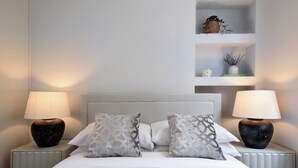 A neatly made bed with two decorative pillows, flanked by two bedside tables with lamps. There are two shelves above the bed, each containing decorative items.