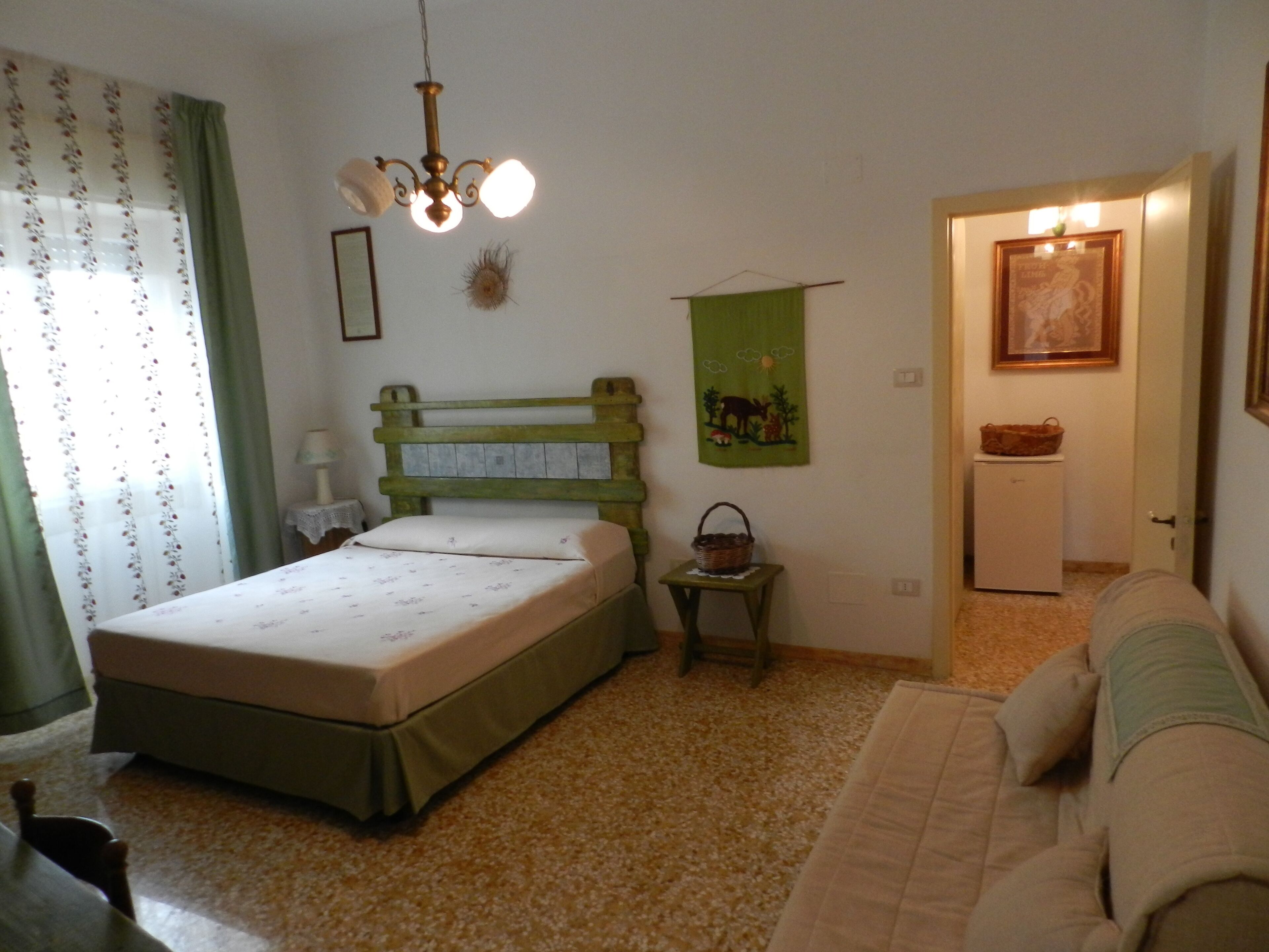 A bedroom with a bed, a green headboard, a small table with a basket, a green wall hanging, a white refrigerator, and a door leading to another room.
