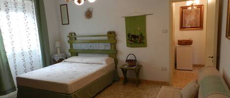 A bedroom with a bed, a green headboard, a small table with a basket, a green wall hanging, a white refrigerator, and a door leading to another room.