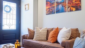 Un salon avec un canapé, des coussins, une table basse et un vase. On y trouve une porte avec un panneau de verre et un petit cadre photo accroché au mur.