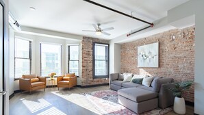 A spacious living room with a large sectional sofa, two armchairs, a coffee table, and a patterned rug. The room features exposed brick walls, large windows with blinds, and a ceiling fan.