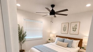 Ein Schlafzimmer mit einem Bett, zwei Nachttischen, einem Deckenventilator, einem Fenster mit Jalousien und einer Topfpflanze.