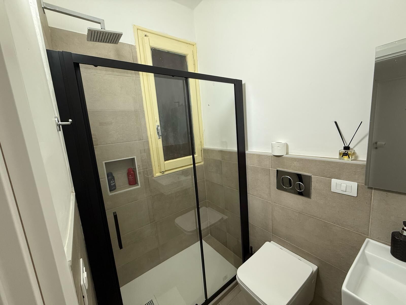 Un baño moderno con ducha de vidrio, inodoro, lavamanos y un estante pequeño con artículos de aseo.