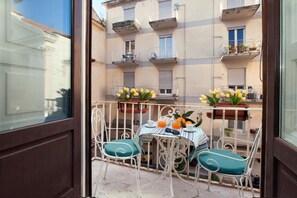 A balcony with a table set for two, featuring a striped tablecloth, two chairs with teal cushions, and a vase of yellow flowers.
