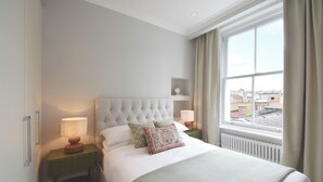 Une chambre à coucher bien rangée, avec un grand lit, deux tables de chevet avec des lampes, une petite étagère et une fenêtre avec des rideaux.