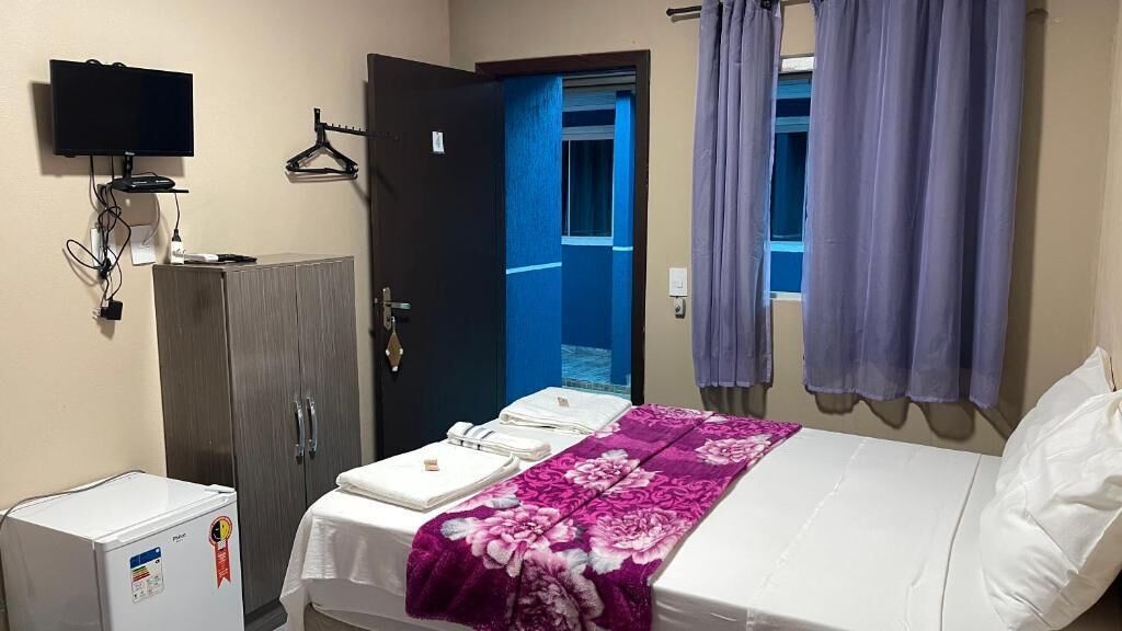 Quarto de hotel com cama, guarda-roupa, televisão e pequeno frigorífico.