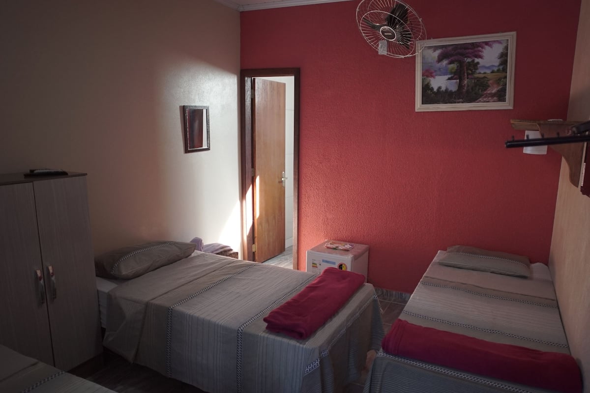 A room with two beds, a wooden wardrobe, a small table with a fan, a painting on the wall, and a door leading to another room.