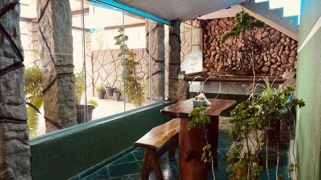 Pátio com paredes de pedra, mesa e banco de madeira, plantas em vasos e vista para o exterior.