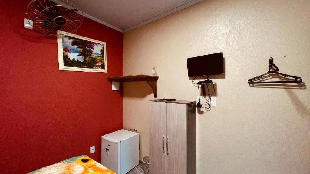 Quarto pequeno com parede vermelha, armário branco, geladeira branca compacta, TV fixada na parede e ventilador de parede.