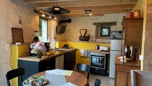 A compact kitchen with a dining area, wooden ceiling beams, a small window, and a refrigerator. - La Grange du Manoir (eco-cottage for 2–4 people) (Le Vieux-Marché)