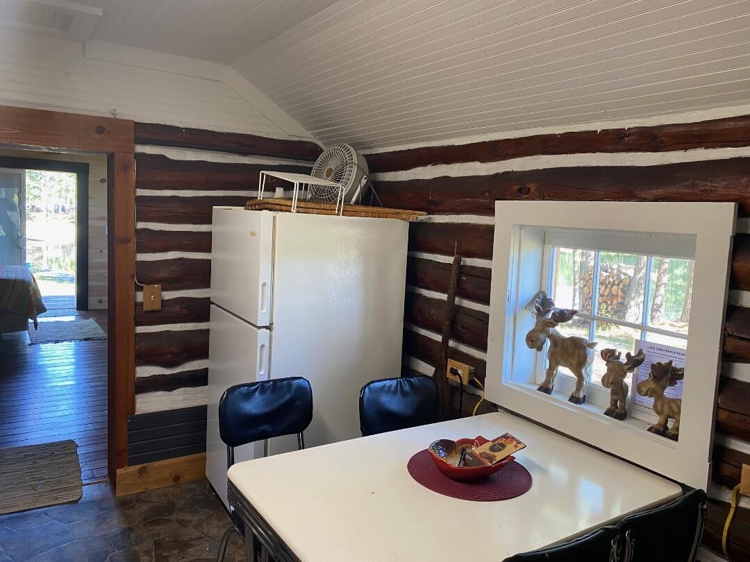 A rustic cabin interior with a wooden log wall, a white refrigerator, two blue chairs, a dining table with a red placemat, and a window with a framed picture of animals.